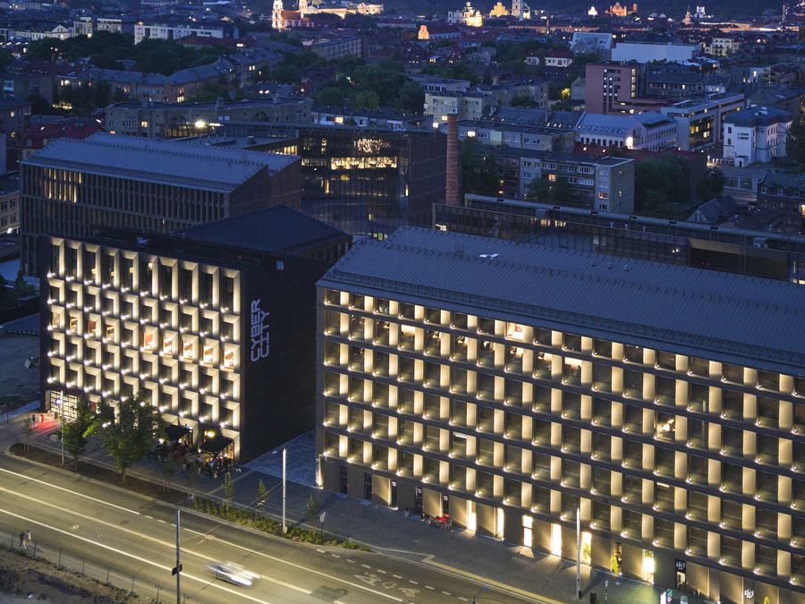 Cyber City in Vilnius at dusk, seen from above: lit office facades by the street, the old factory chimney behind them and the Old Town on the horizon